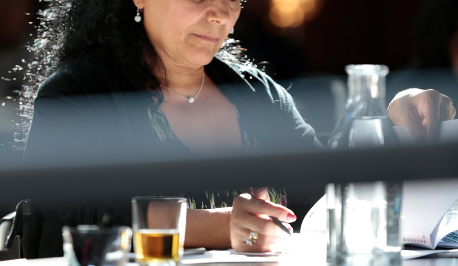 Kvinna med mörkt långt hår, en mörk blus och mönstrad kjol. Sitter vid ett bord och skriver.