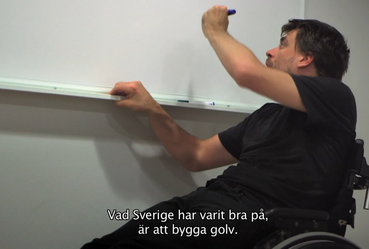 En vit, mörkhårig man i rullstol ska börja rita på en whiteboard. Undertext: Vad Sverige har varit bra på, är att bygga golv.