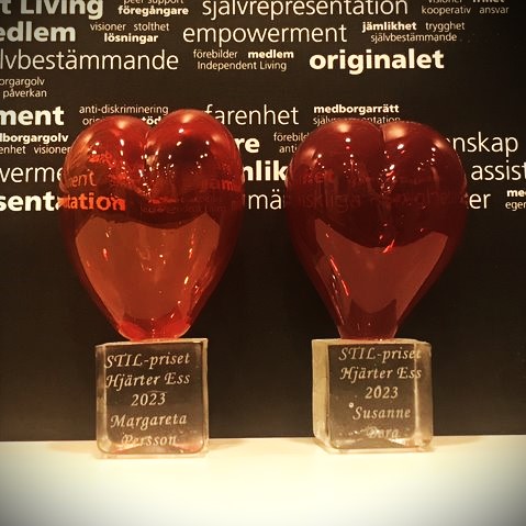 Två röda glashjärtan bredvid varandra. På den ena står STIL-priset Hjärter Ess 2023 Margareta Persson och på den andra STIL-priset Hjärter Ess 2023 Susanne Berg. En svart vägg med vita ord i bakgrunden.