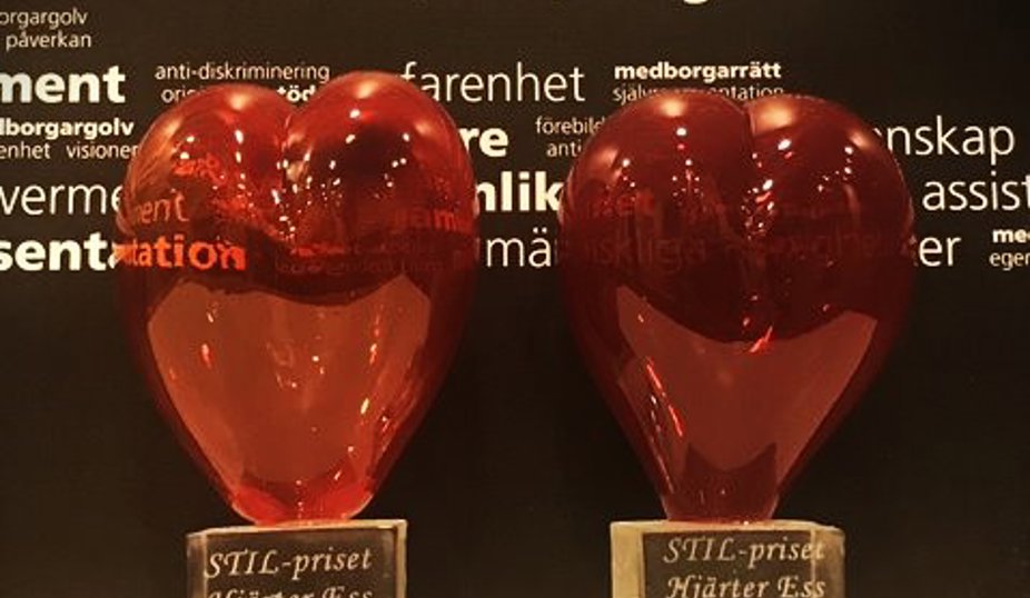 Två röda glashjärtan bredvid varandra. På den ena står STIL-priset Hjärter Ess 2023 Margareta Persson och på den andra STIL-priset Hjärter Ess 2023 Susanne Berg. En svart vägg med vita ord i bakgrunden.
