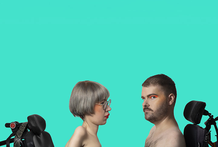 En kvinna och en man sitter mitt emot varandra i varsin permobil. Båda har nakna axlar. Kvinnan har kort grålila hår och glasögon och tittar på mannen. Mannen har mörkt hår, lite skägg och turkos ögonskugga. Han tittar in i kameran. Bakgrunden är turkos.