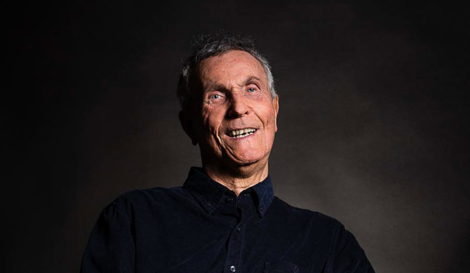 Adolf är en vit äldre man med kort grått hår i svart skjorta och byxor. Han sitter i elrullstol och ler mot kameran. I handen håller han ett munstycke till sin andningsapparat.