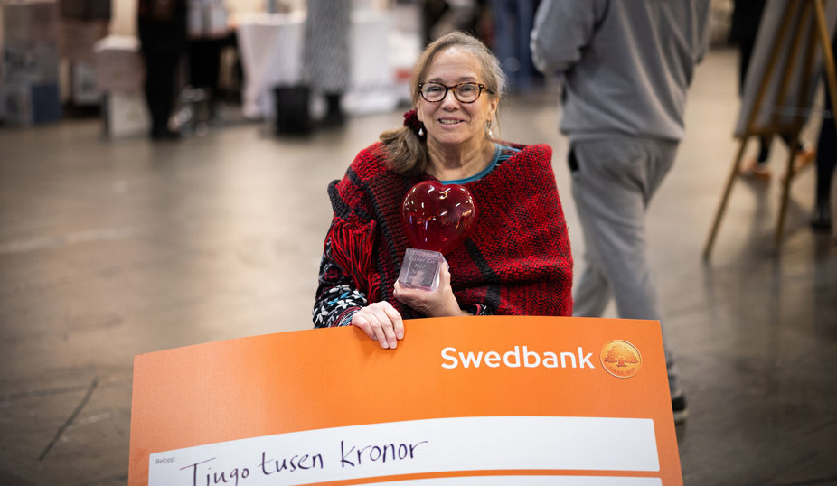En kvinnai rullstol håller ett rött hjärtformat pris och står bakom en stor orange Swedbank-skylt. Personen ler mot kameran i en livlig inomhusmiljö.