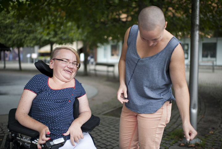 Två personer utomhus; en person i rullstol ler, medan den andra står bredvid och tittar ner med ett leende.