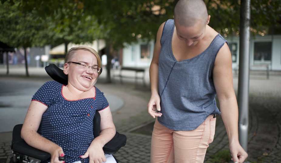 Två personer utomhus; en person i rullstol ler, medan den andra står bredvid och tittar ner med ett leende.