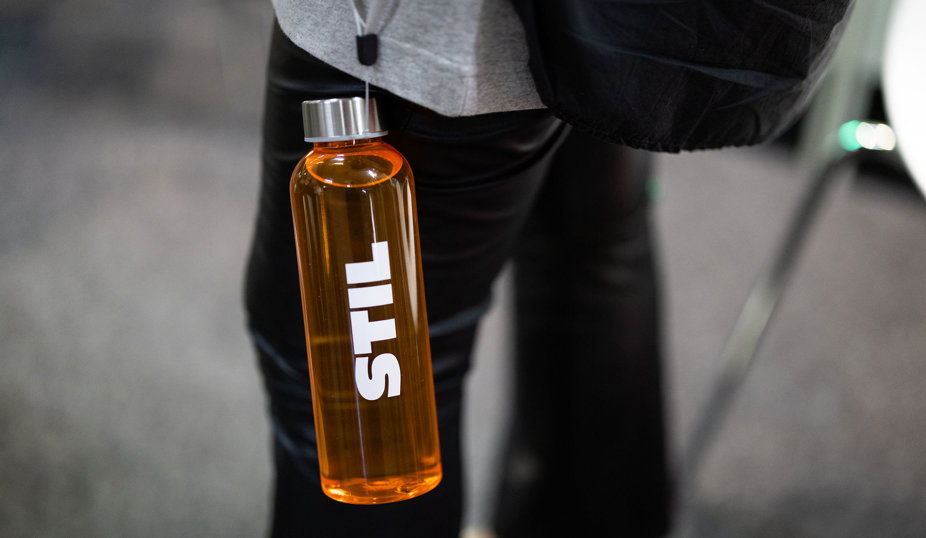 Orange vattenflaska med vit STIL-logotyp som hänger på en person med svarta byxor.