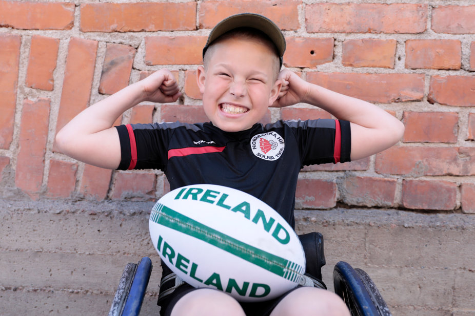 Vit 12-årig kille med ljust hår i rugby-rullstol och rugbyboll i knät.