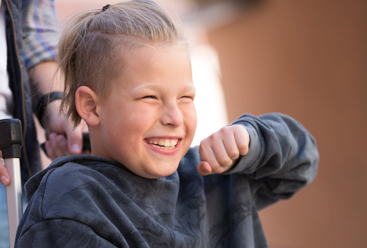 Vit 12-årig kille med ljust hår i rullstol som ser glad ut.