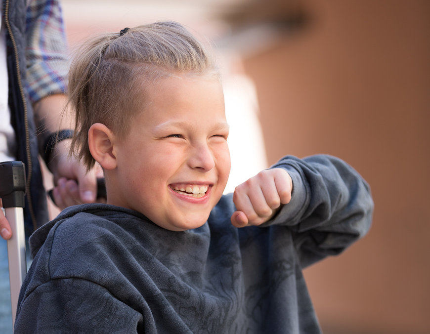 Vit 12-årig kille med ljust hår i rullstol som ser glad ut.