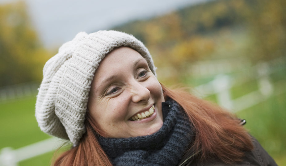 En kvinna med mössa och halsduk ler stort utomhus med ett landskap i bakgrunden.