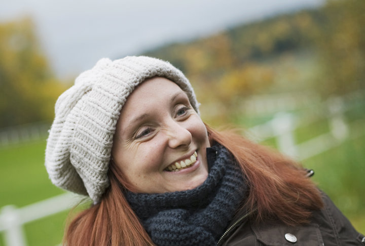 En kvinna med mössa och halsduk ler stort utomhus med ett landskap i bakgrunden.