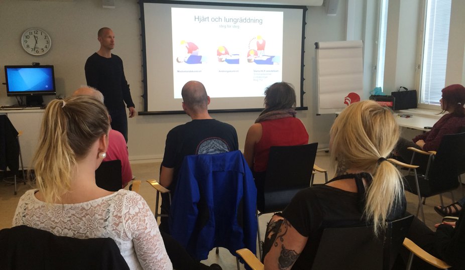 En grupp personer deltar i en utbildning om hjärt- och lungräddning. En man står vid en projektorduk och presenterar instruktioner medan deltagarna sitter på stolar i ett klassrum.