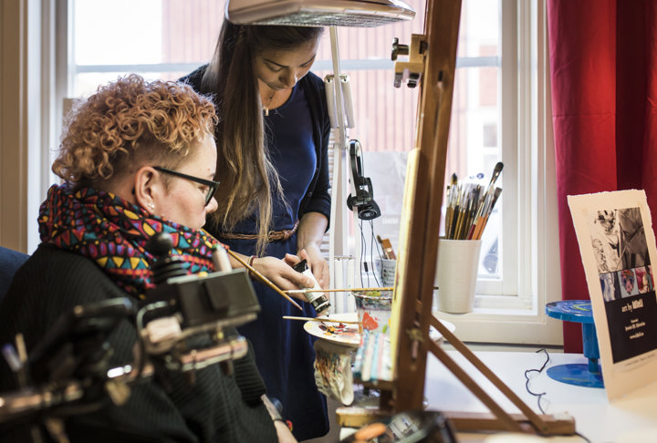 En person målar med munnen. En annan person assisterar med färger.