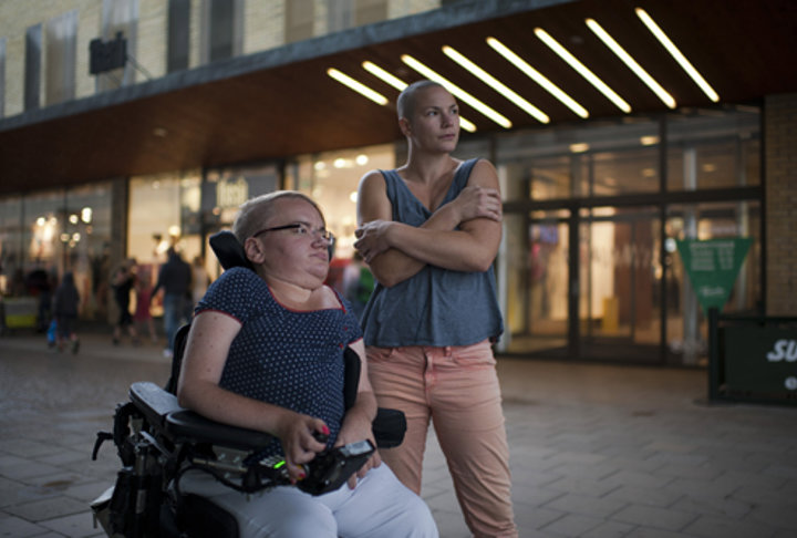 Två personer står utanför en upplyst byggnad. Personen till vänster sitter i en rullstol, och personen till höger står bredvid, lutad med armarna i kors. Båda blickar mot något i fjärran.