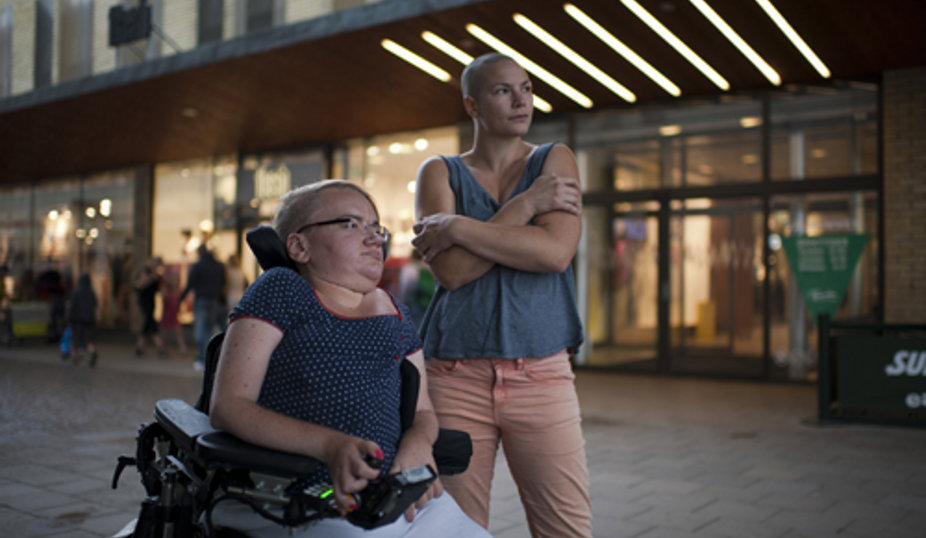 Två personer står utanför en upplyst byggnad. Personen till vänster sitter i en rullstol, och personen till höger står bredvid, lutad med armarna i kors. Båda blickar mot något i fjärran.