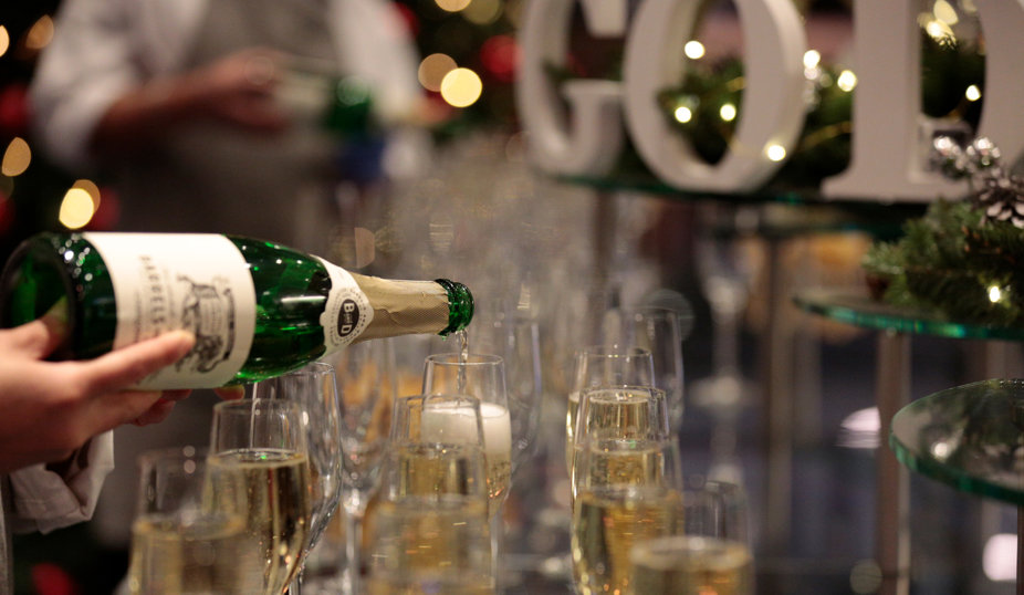 Närbild på någon som häller mousserande vin i flera glas på ett festligt dukat bord med julbelysning i bakgrunden