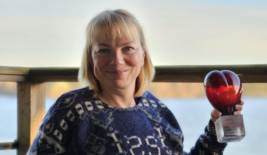 En kvinna i rullstol med en blå mönstrad tröja håller ett rött glashjärta och ler mot kameran.