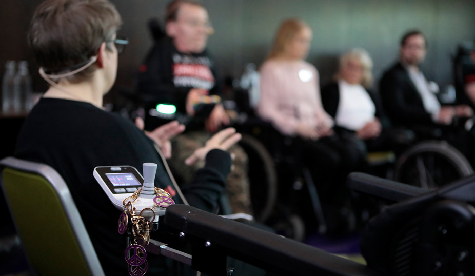 En person i rullstol i förgrunden, omgiven av andra deltagare i ett samtal eller paneldiskussion.