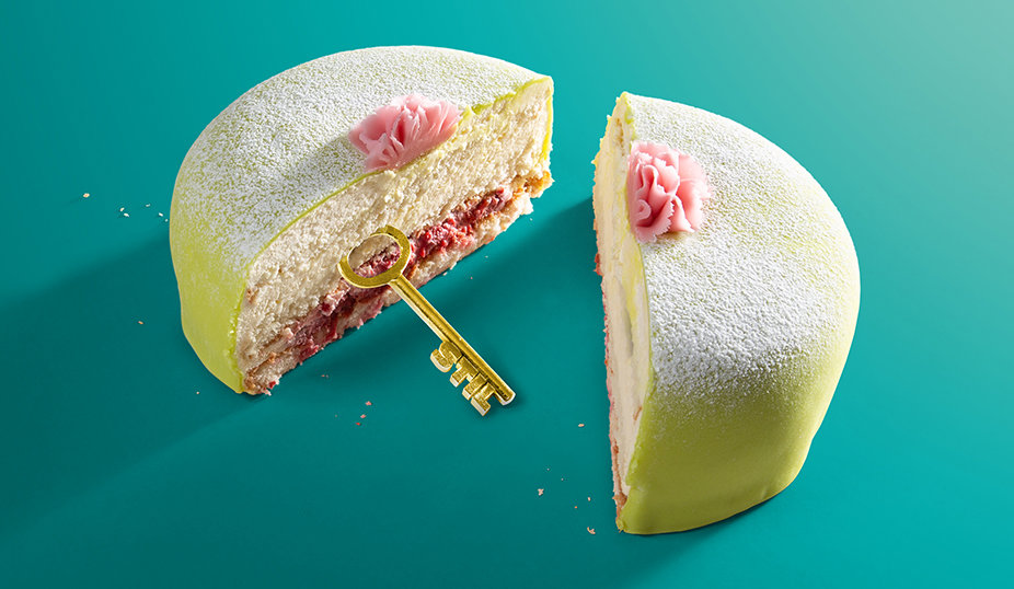 En prinsesstårta med grön marsipan, rosa dekorativa rosor och ett lager fyllning synligt, med en guldnyckel instucken i tårtan, placerad mot en turkos bakgrund.