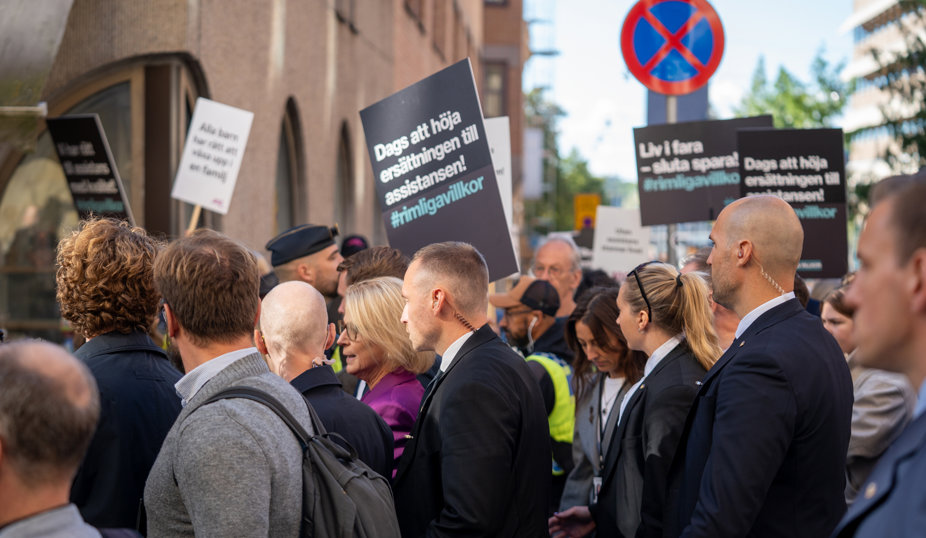 Demonstration med personer som håller skyltar om assistansersättning på en stadsgata.
