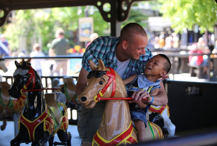 Mörkhyad pojke omkring omkring fyra år, sitter på en häst i karusell. Bredvid står hans pappa.