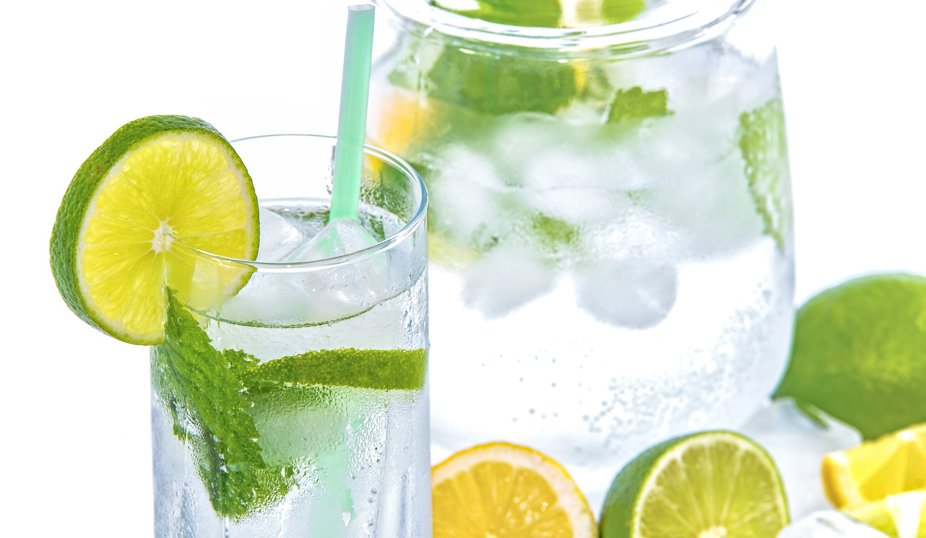 Ett glas och en kanna fyllda med isvatten, lime- och citronskivor samt färska myntablad. Glaset har en grön limeklyfta på kanten och ett sugrör i vattnet.