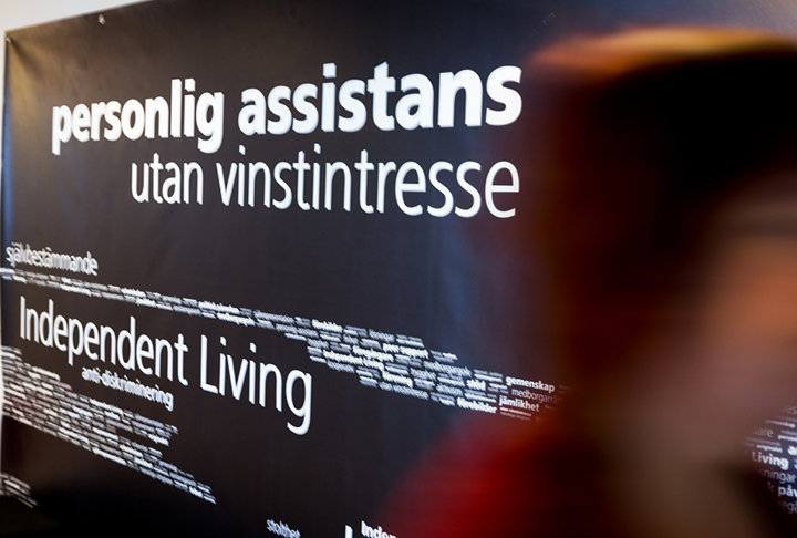 En vägg med texten 'personlig assistans utan vinstintresse' och 'Independent Living' på en mörk bakgrund, där en suddig person passerar i förgrunden