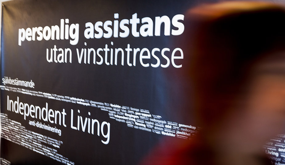 En vägg med texten 'personlig assistans utan vinstintresse' och 'Independent Living' på en mörk bakgrund, där en suddig person passerar i förgrunden