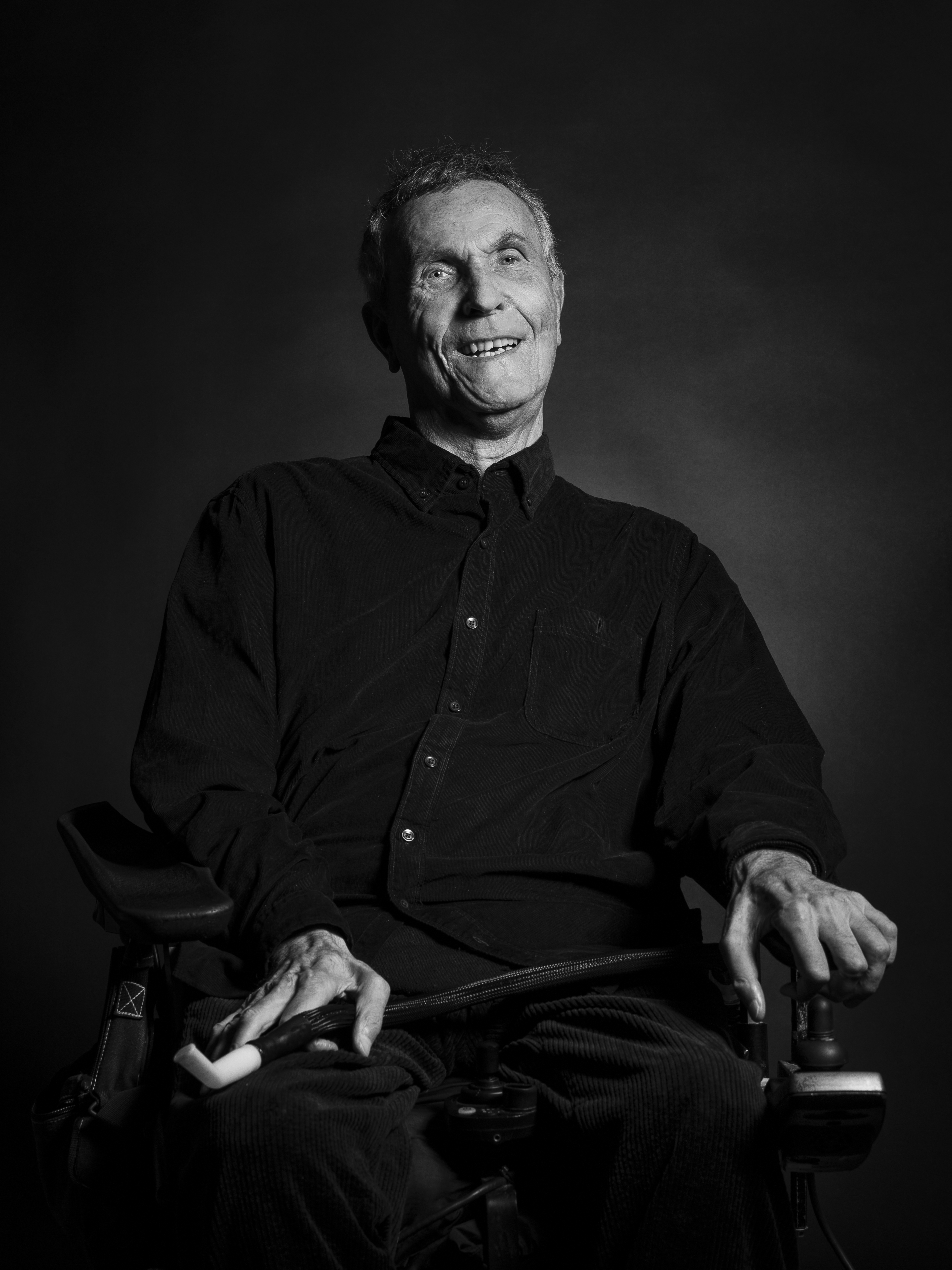 Svartvitt porträtt av en äldre man i rullstol, klädd i en mörk skjorta. Han ler och har ena handen på armstödet och den andra på styrspaken till rullstolen. Bakgrunden är enkel och mörk.