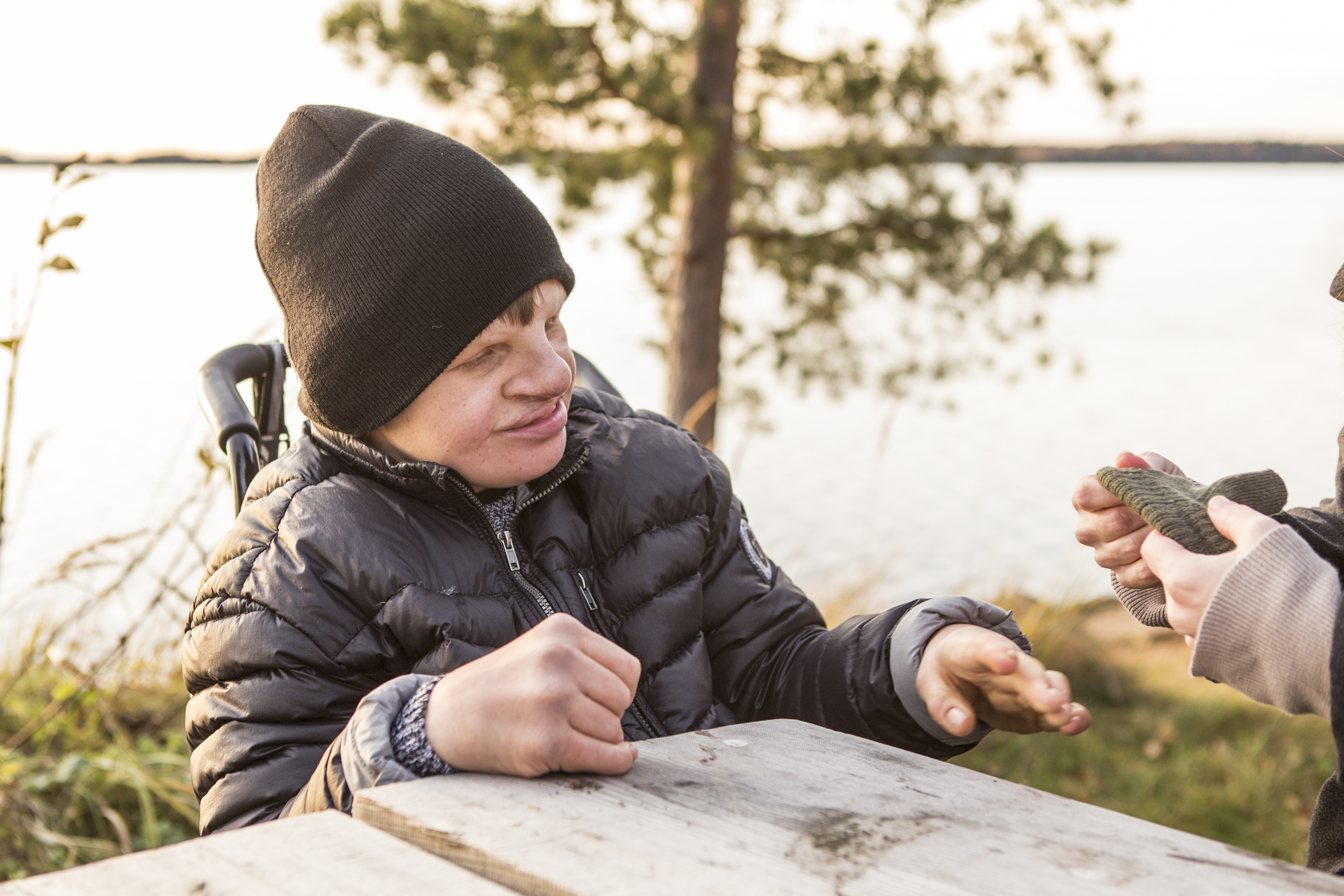 Person i ytterkläder sitter vid ett bord utomhus och sträcker fram handen mot en annan person.