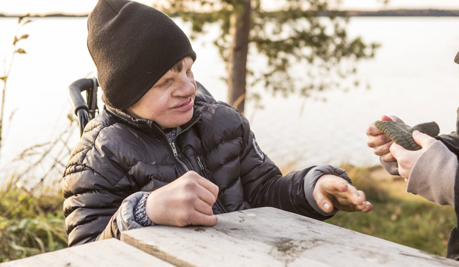 Person i ytterkläder sitter vid ett bord utomhus och sträcker fram handen mot en annan person.