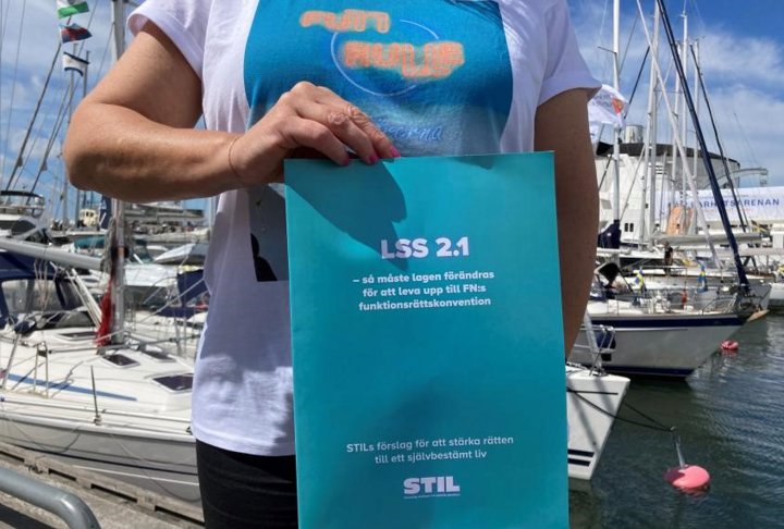 Person håller i STILs rapport LSS  2.1 framför en hamn med segelbåtar.