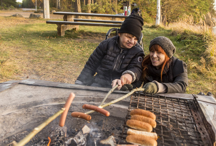 Två personer grillar korv utomhus vid en grillplats. Den ena använder rullstol.
