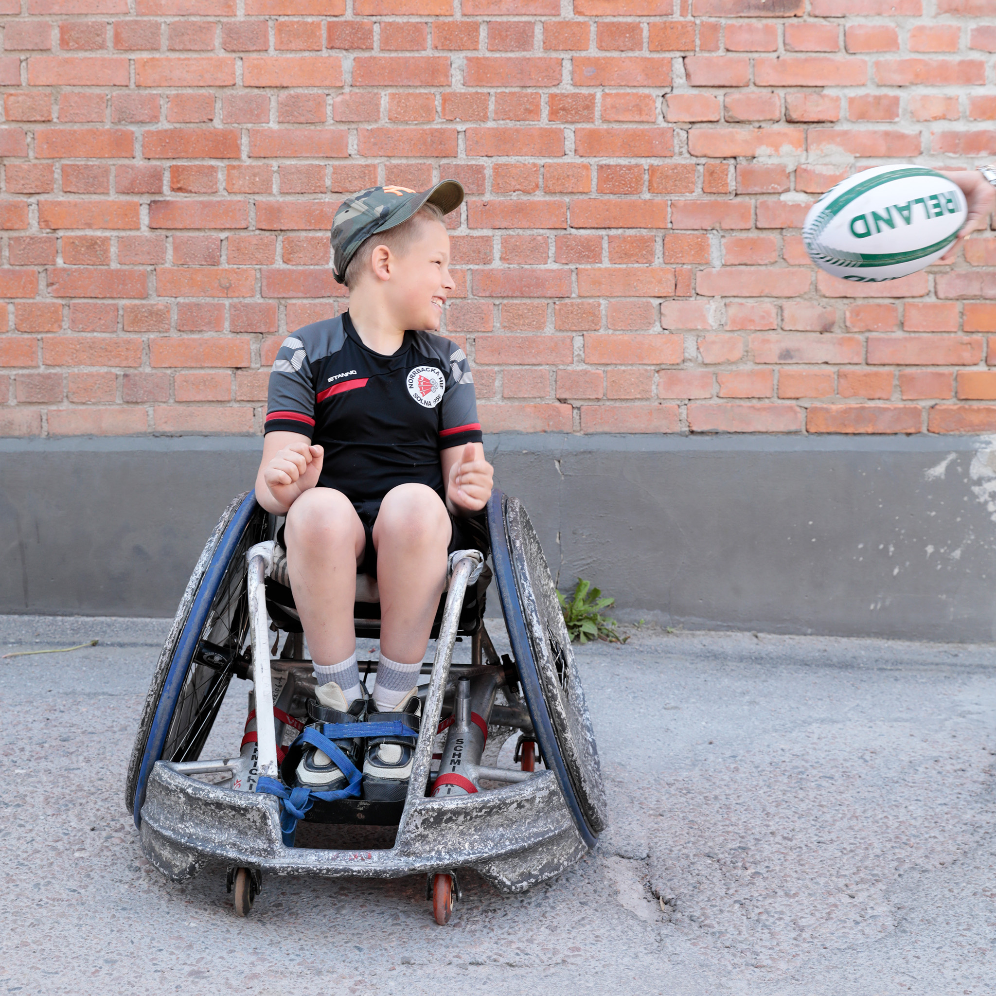 Vit 12-årig kille med ljust hår i rugbyrullstol, tar emot en rugbyboll.