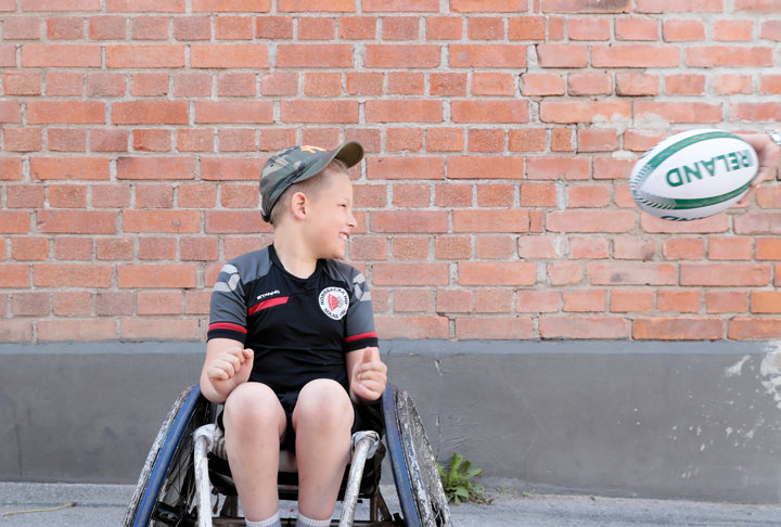 Vit 12-årig kille med ljust hår i rugbyrullstol, tar emot en rugbyboll.