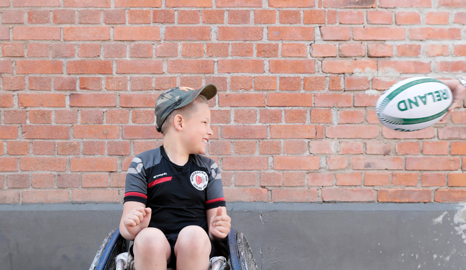 Vit 12-årig kille med ljust hår i rugbyrullstol, tar emot en rugbyboll.