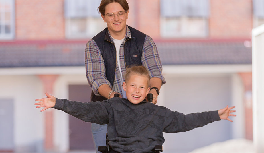 En glad pojke i rullstol sträcker ut armarna medan en vuxen person bakom honom skjuter stolen, i en ljus och utomhusmiljö