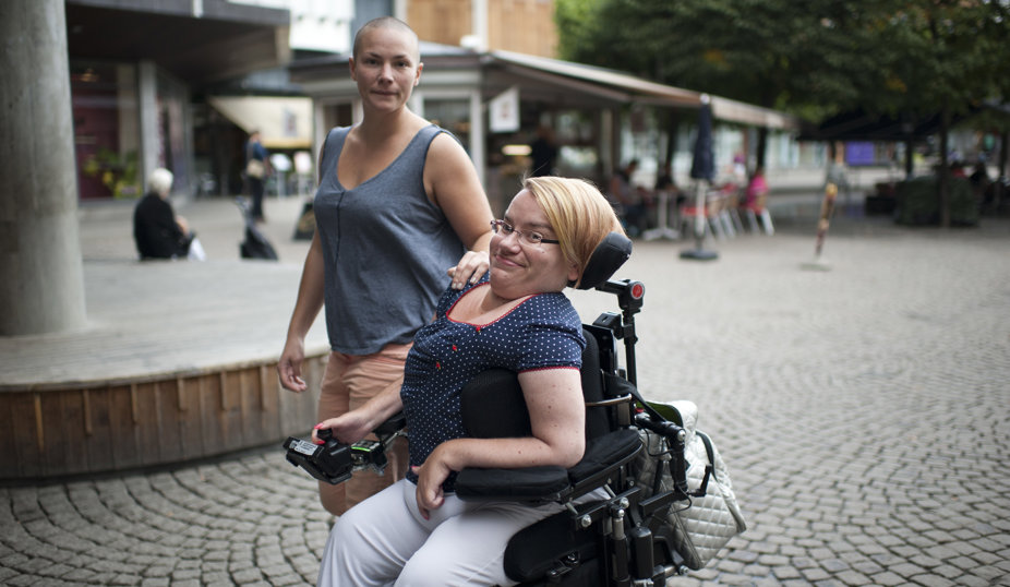 vå personer utomhus; en person sitter i en rullstol och ler mot kameran, medan den andra står bredvid med en hand på rullstolens ryggstöd och tittar in i kameran.